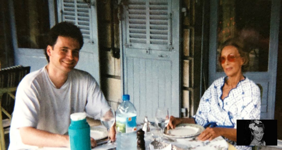 Fotografia colorida de uma mesa de almoço diante de venezianas azuis: um jovem à esquerda, Aimée de Heeren à direita, de óculos escuros